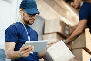 Delivery workers handling packages in a warehouse, one checking shipment details on a tablet device.