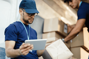 Delivery workers handling packages in a warehouse, one checking shipment details on a tablet device.