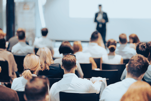 Audience attending a business presentation with a speaker presenting insights on a large screen.